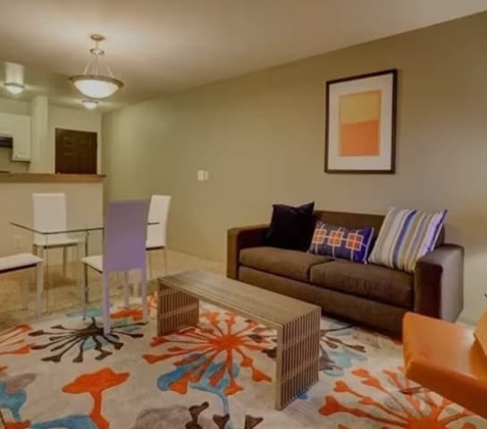 Modern living room at The View on 26th Boulder with a brown sofa, colorful pillows, abstract art, and a glass dining table with white chairs. A vibrant rug in orange and blue floral patterns brightens the space, with a small kitchen in the background.