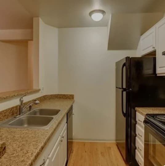 A small kitchen at The View on 26th Boulder features granite countertops, a double sink, white cabinets, a black refrigerator, and stove. Light wood floors and a bright ceiling light complete the space.