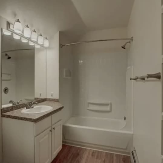 A bathroom at The View on 26th Boulder features a white bathtub and shower, a single sink vanity with granite countertop, a large mirror, overhead lights, and wood-patterned flooring.