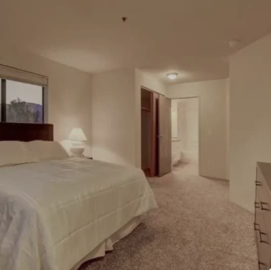 A cozy bedroom at The View on 26th Boulder features beige walls, carpeted floor, a neatly made bed with white bedding, bedside table with lamp, a window, dresser, and an open doorway leading to a bathroom.