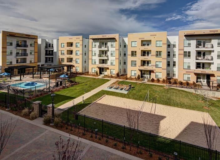 The U Club on 28th Boulder features a courtyard with four modern beige and white buildings, a large green lawn, outdoor pool with blue umbrellas, volleyball sand court, and a shaded pergola area under a partly cloudy sky.