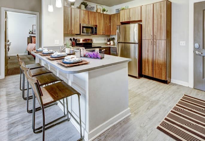 Modern kitchen at U Club on 28th Boulder with wooden cabinets, stainless steel appliances, and a long island with four stools. The island is set for dining, and a striped rug decorates the light wood floor near the entrance.