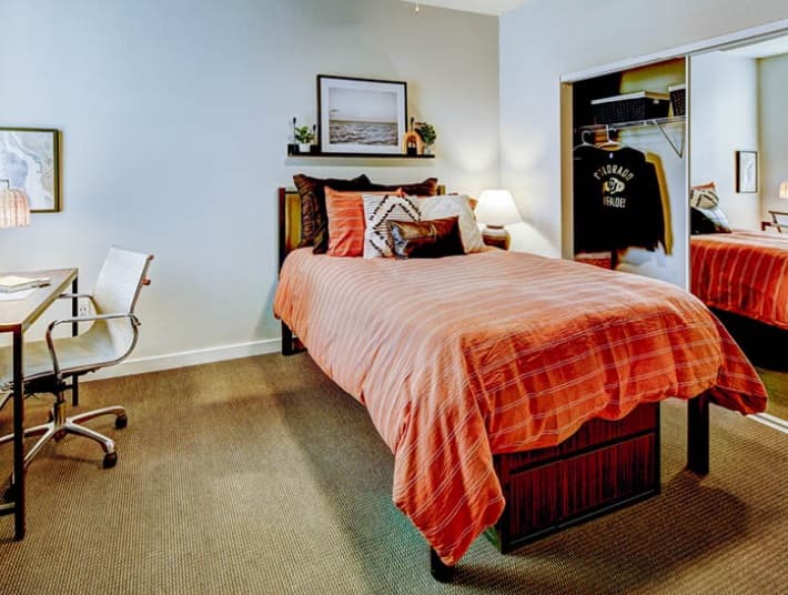 A neatly organized U Club on 28th Boulder bedroom with a bed covered in a red striped comforter, a desk and chair on the left, wall art above the bed, and an open closet with clothes and a mirror on the right.