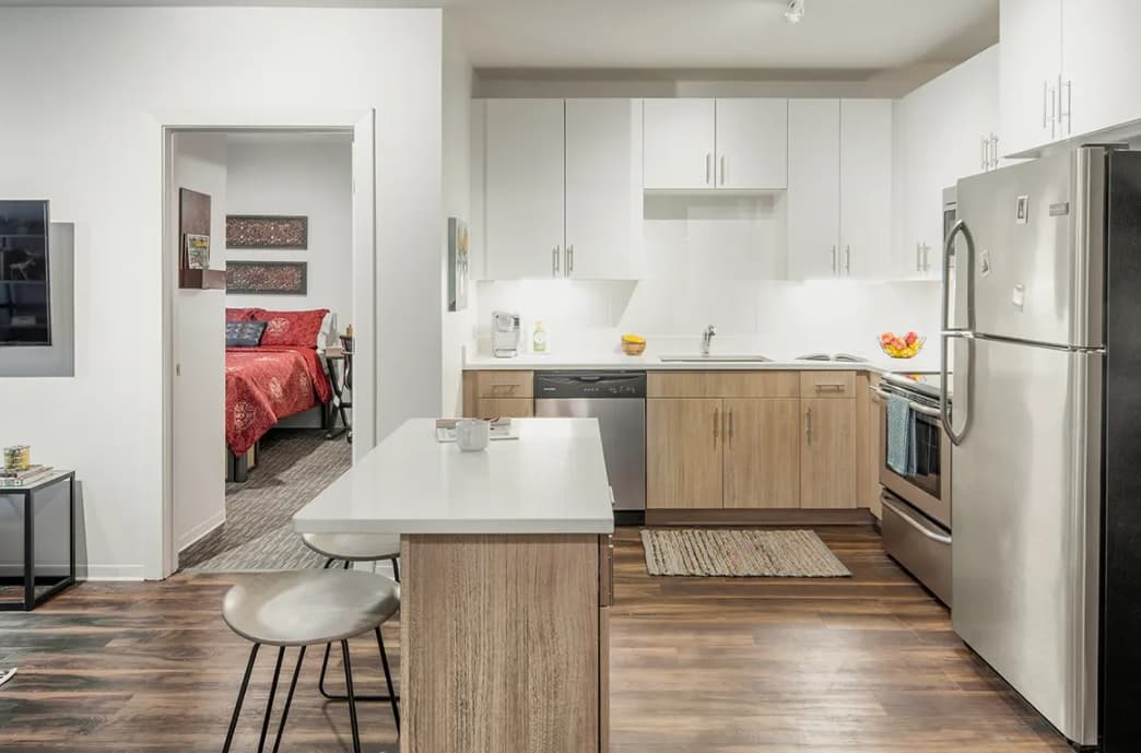 Modern kitchen with white cabinets, stainless steel appliances, and an island with two stools. An uncommon Fort Collins touch: a bedroom with a red bedspread is visible through the open doorway. The wood floor and ample light enhance the space.