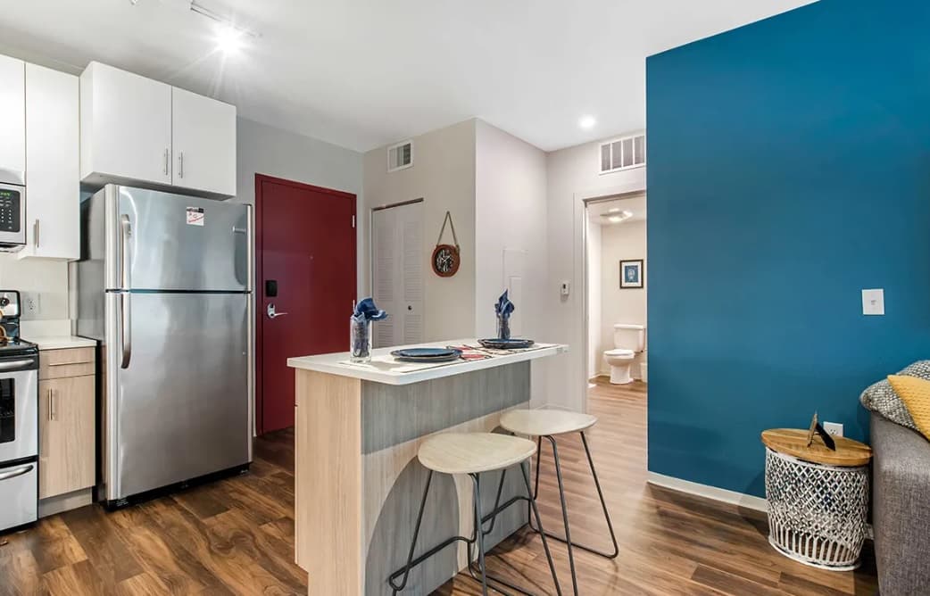 Modern kitchen with stainless steel appliances, a small island with two stools, wood flooring, blue accent wall, and a view into a bathroom. Discover Uncommon Fort Collins style in the decorative items on the island and basket near the couch.