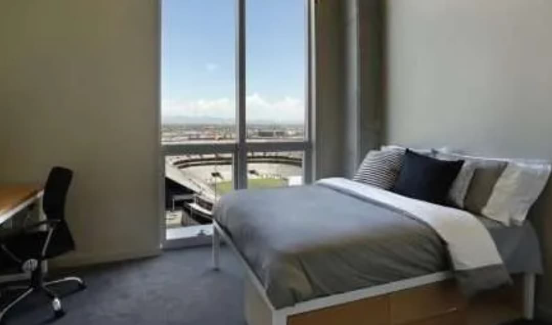 A modern bedroom at University House Tempe features a neatly made bed, a desk with a chair, and large floor-to-ceiling windows offering views of the stadium and distant mountains under a blue sky.