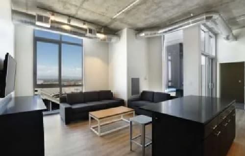 Modern apartment living room at University House Tempe with industrial-style ceiling, exposed ductwork, large windows showcasing a city view, two black sofas, a wooden coffee table, and a kitchen island with a metal stool.