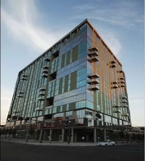 A modern, multi-story glass building with geometric balconies stands on a city corner. The sky is clear, and sunlight reflects off the windows of University House Tempe. A white car is parked near the entrance.