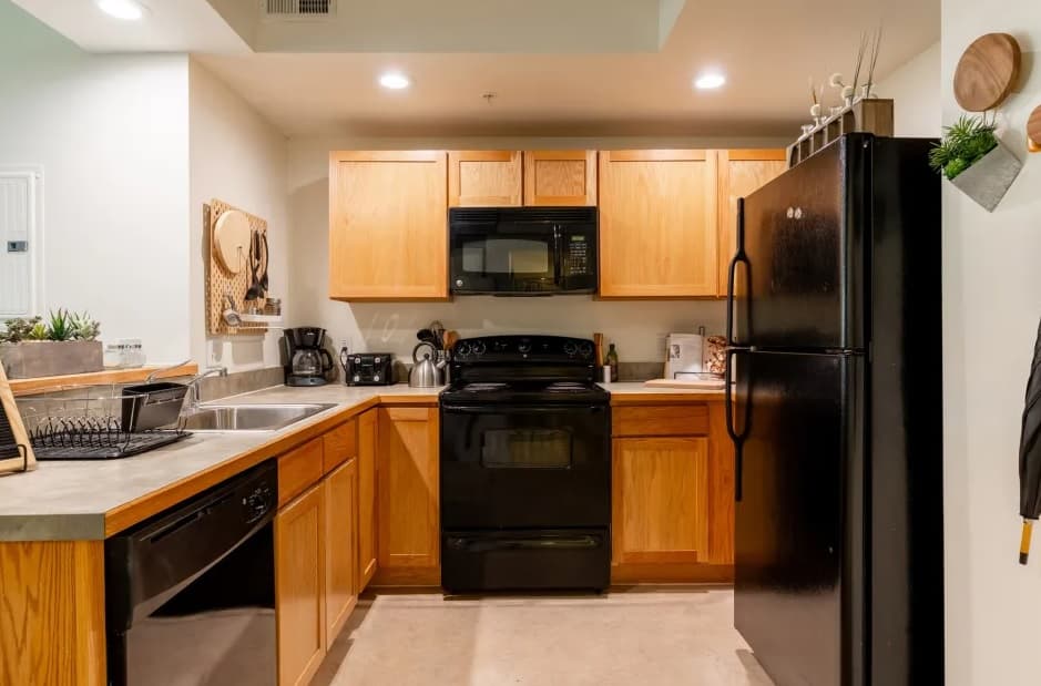 A modern kitchen at University House Denver features wooden cabinets, black appliances including a refrigerator, stove, dishwasher, and microwave, a stainless steel sink, and various small kitchen items on the counters.