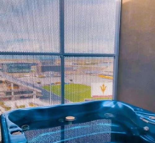 A blue hot tub on a balcony at University House Tempe overlooks a sports field with an ASU logo, seen through a mesh screen with buildings and a clear sky in the background.