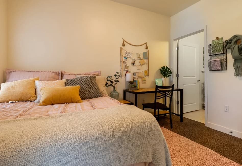 A cozy University house Denver bedroom with a neatly made bed, pastel pillows, a desk and chair, bulletin board, and soft lighting. A plant sits on the desk near a door leading to another room. The decor is tidy and inviting.