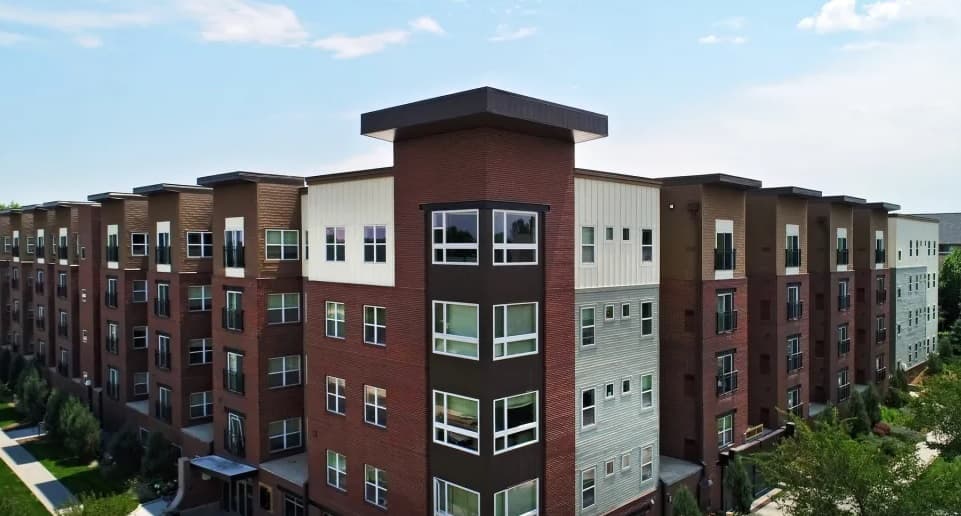A modern four-story apartment building near University House Denver, with a mix of brick and light-colored siding, featuring many windows, balconies, and lush green landscaping under a partly cloudy sky.