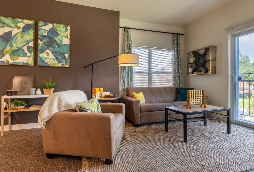 A cozy living room in a University house Denver features brown sofas, a black coffee table, a standing lamp, and wall art. Large windows with curtains fill the space with natural light, while plants sit on a console table against a brown accent wall.
