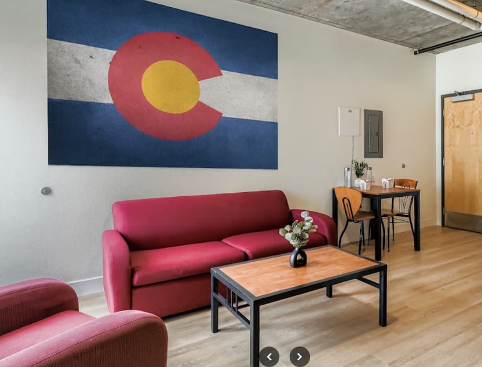 A living room at University Lofts with a red sofa and armchair, a wooden coffee table, a small dining table for two, and a large Colorado state flag hanging on the wall above the sofa.