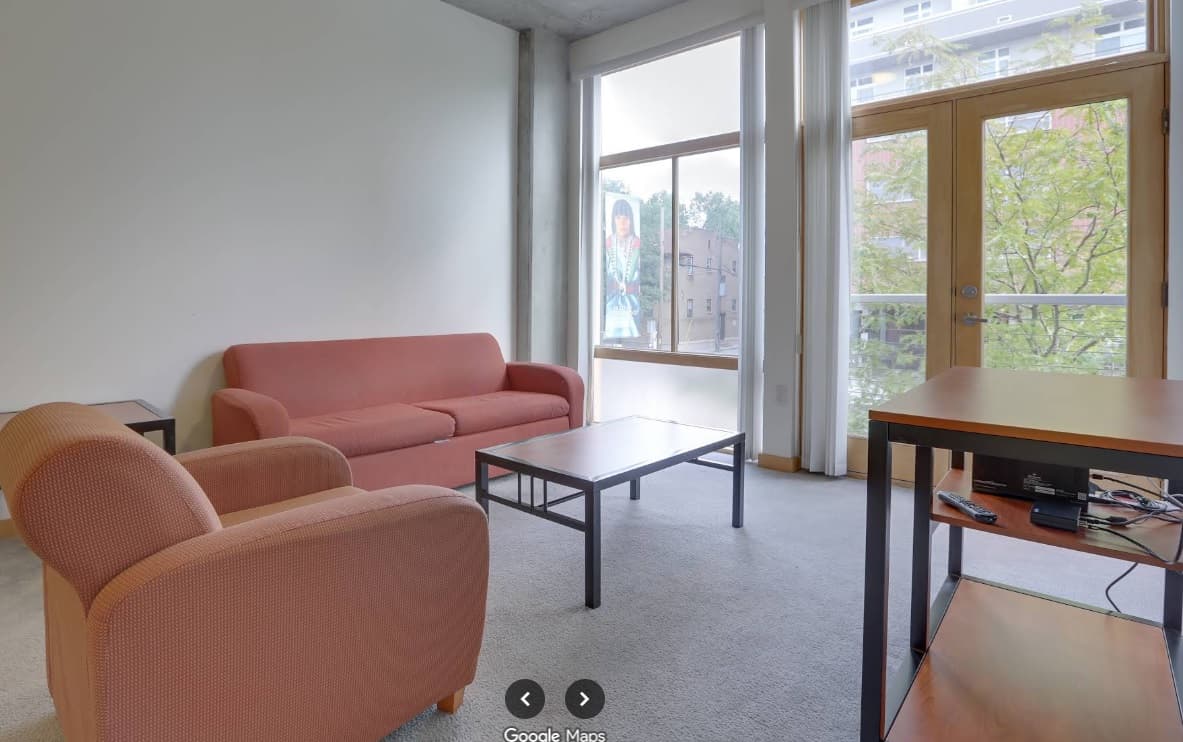 A modern living room at University Lofts features a pink sofa and armchair, black coffee table, wooden desk with electronics, large windows, and a glass door opening to a balcony with trees visible outside.