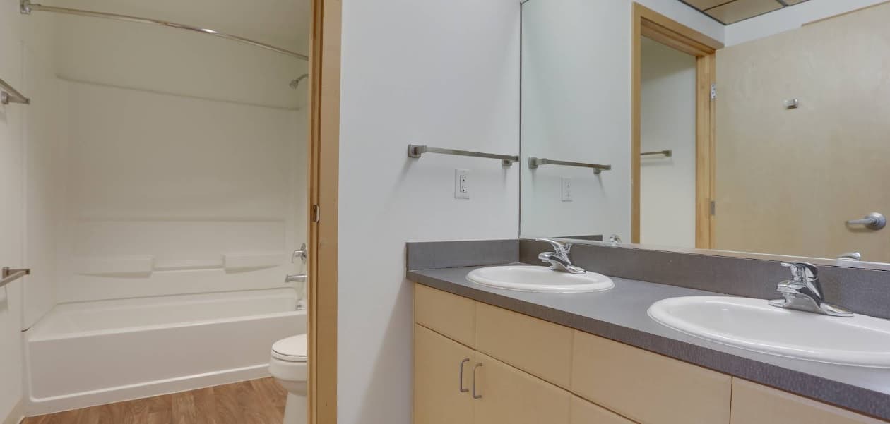 A modern bathroom at University Lofts features a double-sink vanity, light wood cabinets, a large mirror, and a white bathtub with shower curtain rod. The wood-patterned floor adds warmth, and a door is visible in the background.