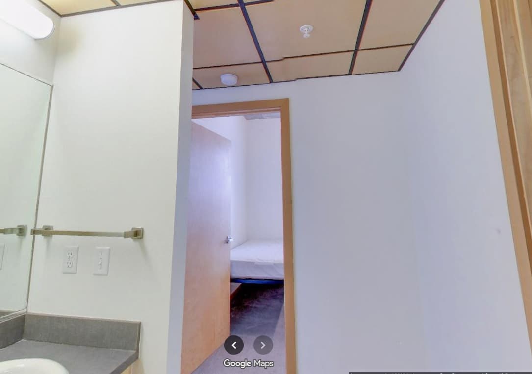 View from a bathroom with a sink and mirror, looking through an open door into a bedroom at University Lofts, featuring a made bed, white walls, and a ceiling with a grid pattern of square tiles.