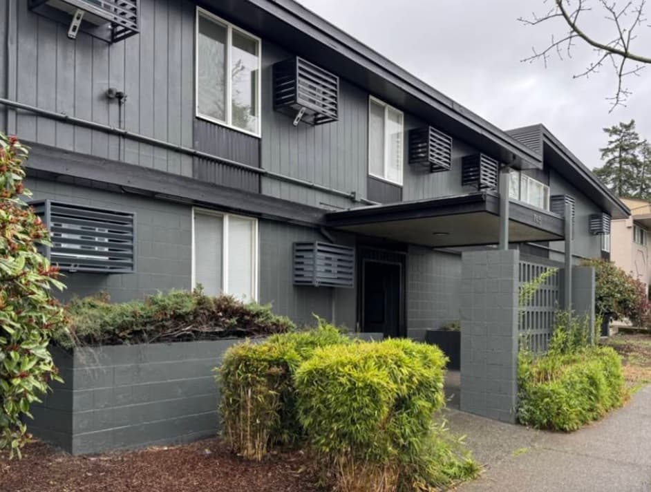 A modern, two-story gray apartment building, University Manor features large windows with metal shades, surrounded by shrubs and greenery under an overcast sky.