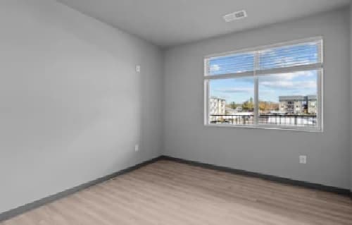 An empty room with light gray walls, wood flooring, and a large window letting in natural light. The window offers a view of neighboring buildings and the Pacific Highway under a partly cloudy sky.