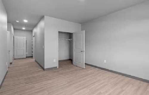 Empty room with light-colored wood flooring, gray walls, and white doors at Union at Pacific Highway. An open closet with a wire shelf is visible, and two closed doors are in the background along a well-lit hallway.