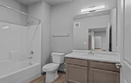 A modern bathroom at Union at Pacific Highway features a white bathtub and shower, toilet, and a vanity with brown cabinets. Light gray walls and wood-look flooring complete the elegant space.