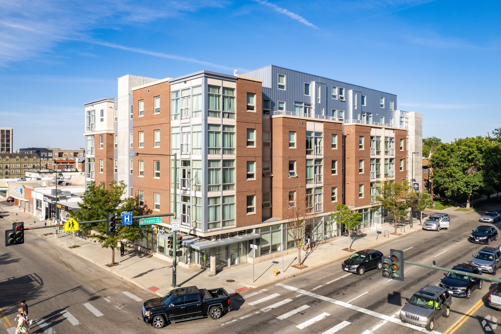 University Lofts, a modern, multi-story apartment building with a brick and glass exterior, stands at a busy street intersection, surrounded by shops, vehicles, pedestrians, and green trees under a clear blue sky.