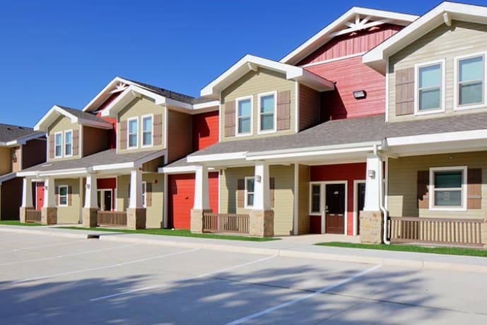 Row of modern, two-story U Club Townhomes with beige and red facades, white trim, small front porches, and empty parking spaces—ideal student housing near Marion Pugh under a clear blue sky.