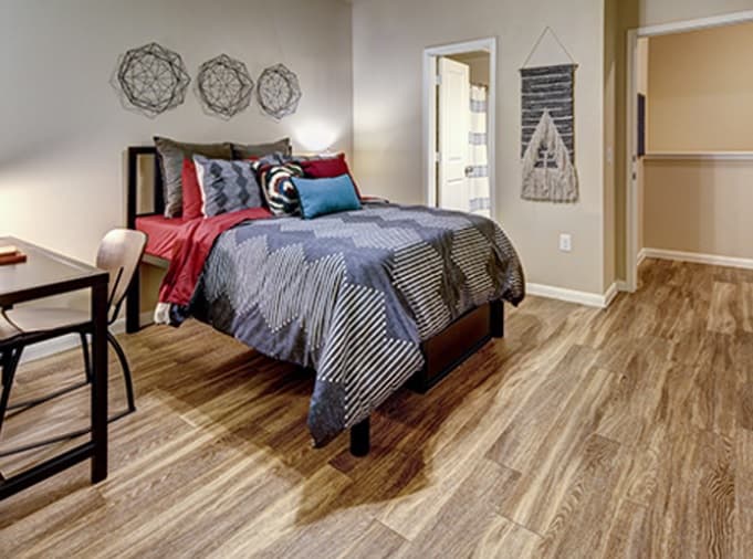 A modern bedroom in U Club Townhomes, ideal for student housing, with a neatly made bed featuring blue and red bedding, geometric wall art above the headboard, a small desk and chair, wood-style flooring, neutral walls, and a tapestry near an open door.