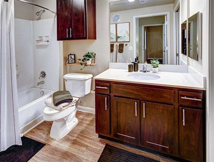 A modern bathroom at U Club Townhomes features dark wood cabinets, a white sink, a toilet with towels on top, a bathtub with shower and white curtain, wall art, decorative vanity items, and stylish wood-style flooring—ideal student housing near Marion Pugh.