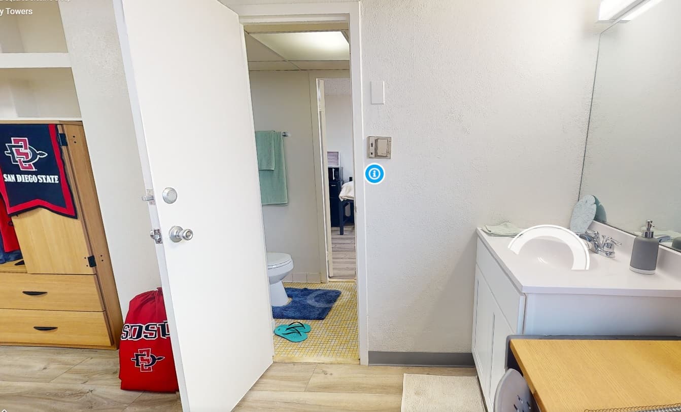 A South Tower dorm room in South Campus Plaza features SDSU decor, a bed and dresser, an open door to a bathroom with towels, and a vanity with sink and mirror. A red SDSU laundry bag rests on the floor—perfect campus housing comfort.