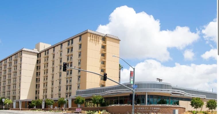 A tall, beige campus housing building known as South Tower stands next to a modern, rounded entrance labeled University Towers under a bright blue sky with scattered clouds. A traffic light and street signs are visible in front.