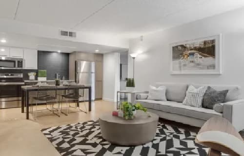 Modern living room at University Valley Apartments Tempe with a gray sofa, patterned rug, round coffee table, wall art, and an open kitchen featuring stainless steel appliances and a dining table with chairs in the background.