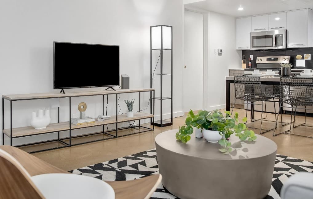 Modern living room at University Valley Apartments Tempe with a TV on a metal and wood stand, geometric rug, round coffee table with green plants, plus a view of the dining area and kitchen with metal chairs and stainless steel appliances.