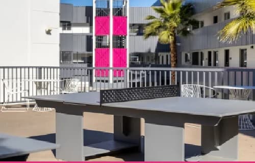 A gray outdoor ping pong table sits on a sunny patio at University Valley Apartments Tempe, surrounded by metal chairs, palm trees, and a modern building with bright pink elevator doors in the background.