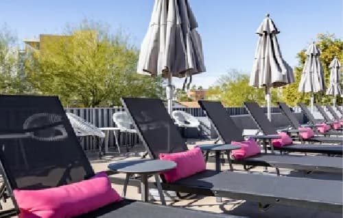 Several black lounge chairs with pink cushions are lined up beside small round tables and closed gray umbrellas on a sunny outdoor patio at University Valley Apartments Tempe, with trees and a fence in the background.