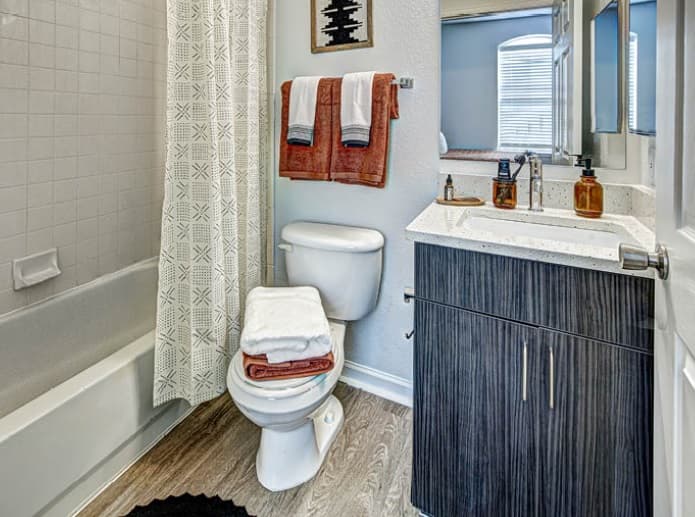 Modern bathroom at University Village at Boulder Creek Boulder with a bathtub and shower curtain, toilet with folded towels on top, sleek vanity with soap and lotion dispensers, mirror, wood-style flooring, and two orange towels hanging on the wall.