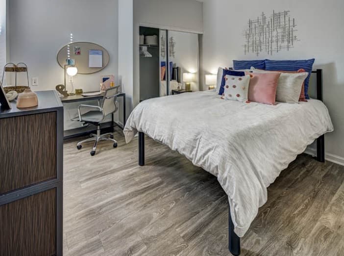 A modern bedroom at University Village at Boulder Creek Boulder features a bed with decorative pillows, a desk with chair and lit lamp, mirror, closet with sliding doors, wood-style flooring, and minimalist decor.