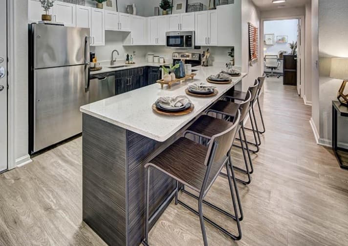 Modern kitchen at University Village at Boulder Creek Boulder features white cabinets, stainless steel appliances, and a large island with four bar stools. The island is set with plates and napkins, and the kitchen opens to a hallway leading to another room.