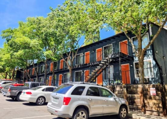 A row of two-story apartments at Elevation at the U Apartments Reno features dark exteriors and vibrant orange doors, shaded by green trees. Several cars are parked in front of the building on a sunny day.