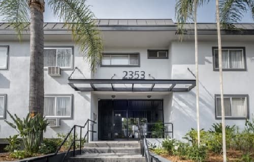 A white two-story apartment building, California Suites, with the number 2353 above the entrance, flanked by palm trees and greenery; concrete steps lead to a glass door with a black metal awning.