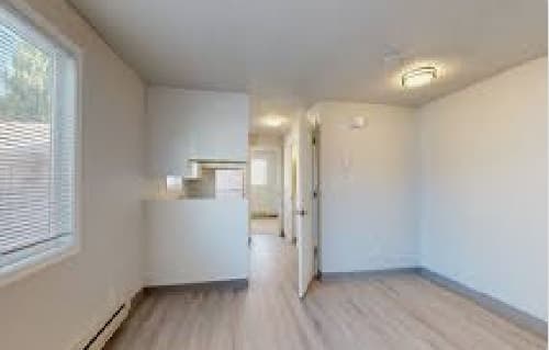 A small, unfurnished apartment room with light-colored walls, wood-style flooring, a window with blinds, and a ceiling light fixture is situated in the vicinity of an open door leading to another area.