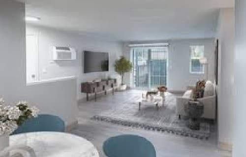 A modern, bright living room with light grey walls and floors, a sofa, coffee table, and TV on the wall. In the vicinity of a large window with blinds sits a green plant. A small dining area is partially visible in the foreground.