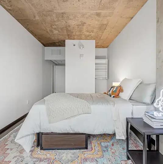 A modern bedroom with a concrete ceiling, white walls, a neatly made bed with light bedding and a textured throw overlooks the stunning vista of Denver, Colorado, complemented by a colorful rug, small bedside table with books, and minimal decor.