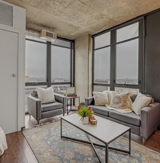 Modern living room with large floor-to-ceiling windows offering a stunning vista of Denver, Colorado. Two gray sofas with cushions, a white coffee table, a patterned rug, and city views complement the concrete ceilings and dark accents.
