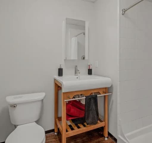 A bathroom with a white toilet, a white sink on a wooden stand with towels and toiletries, a rectangular mirror above the sink, and a shower with white tiles and a curtain rod, reminiscent of modern homes in Vista Denver Colorado.
