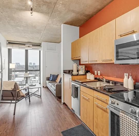Modern studio apartment with wood floors, an orange accent wall, and light wood cabinets. Kitchen has stainless steel appliances. Living space features large windows with a stunning Vista of Denver, Colorado city buildings. Contemporary and cozy decor.