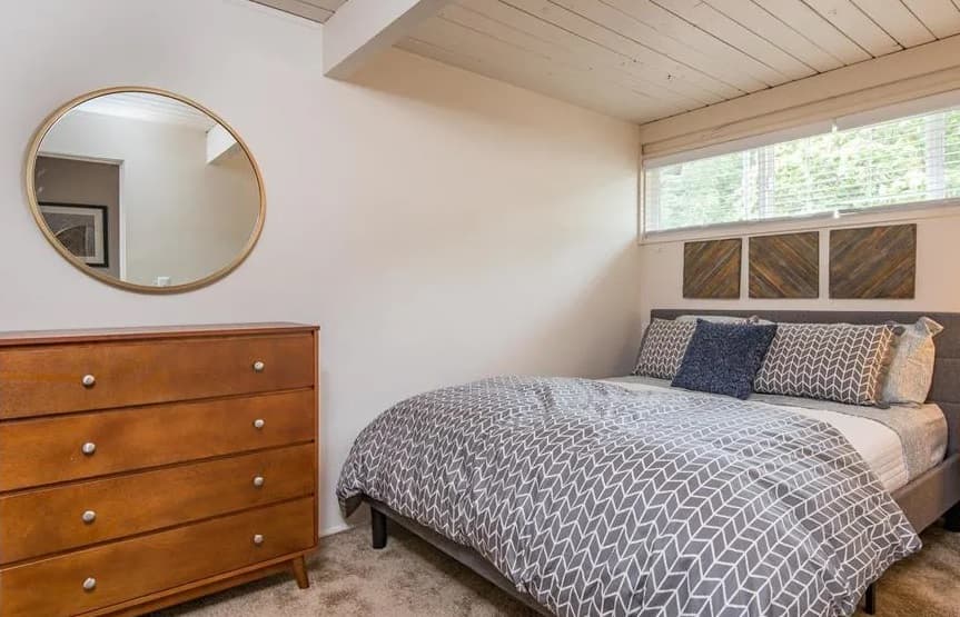 A cozy bedroom at Witham Hill Oaks Apartments features a wooden dresser with a round mirror, a bed with gray-patterned bedding and blue accent pillows, and windows with blinds above the bed letting in natural light.