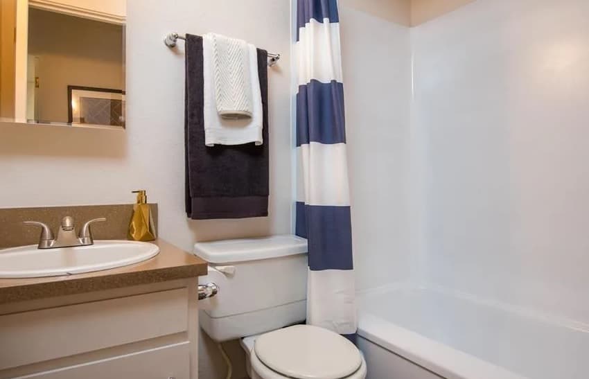 A small bathroom at Witham Hill Oaks Apartments features a white sink, silver faucet, brown countertop, and toilet. Above the toilet are two towels on a rack. A blue and white striped shower curtain covers the bathtub.