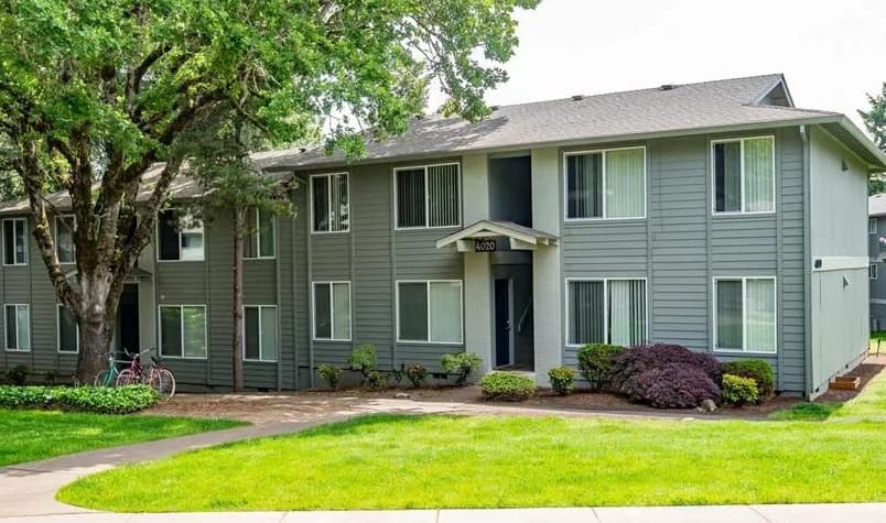 A two-story gray apartment building with white trim, large windows, and a covered entrance sits among green grass, shrubs, and trees at Witham Hill Oaks Apartments. Two bicycles are parked near the front.