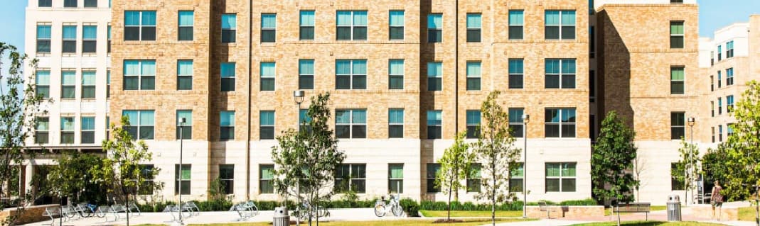 A large, modern brick building with many windows, surrounded by green trees and a paved walkway. Several bicycles are parked outside White Creek Apartments, and the sky is clear and sunny.